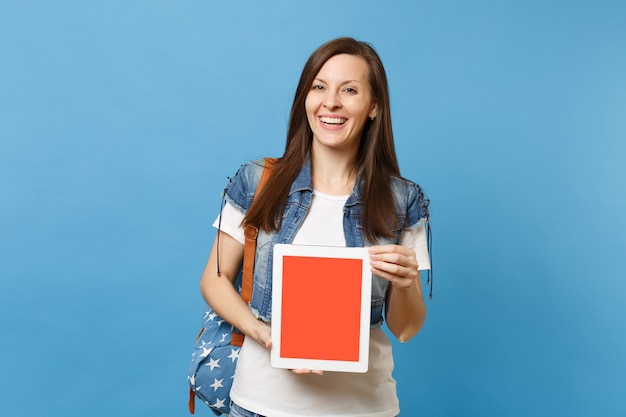 Jonge lachende vrouw student in denim kleding met rugzak met tablet pc-computer met leeg zwart leeg scherm geïsoleerd op blauwe achtergrond. Onderwijs op de universiteit. Kopieer ruimte voor advertentie.