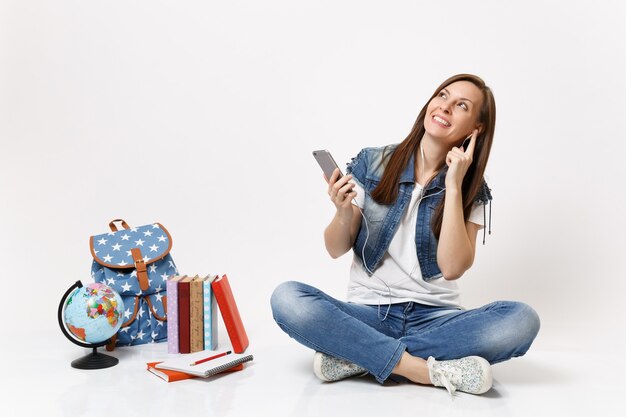 Jonge lachende vrouw student met koptelefoon opzoeken van muziek luisteren met mobiele telefoon zitten in de buurt van globe rugzak boeken geïsoleerd