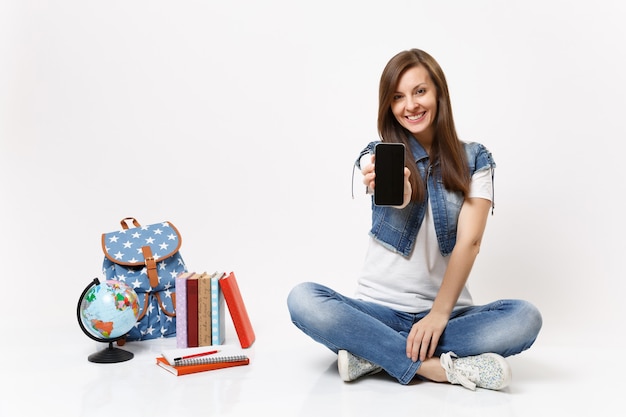 Jonge lachende vrouw student met mobiele telefoon met leeg zwart leeg scherm, zittend in de buurt van globe, rugzak, schoolboeken geïsoleerd