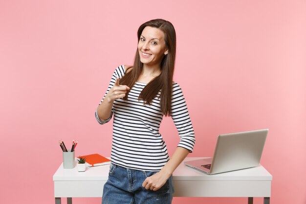 Jonge lachende vrouw wijsvinger wijzend op camerawerk en permanent in de buurt van wit bureau met laptop geïsoleerd op pastel roze achtergrond. Prestatie zakelijke carrière concept. Kopieer ruimte voor advertentie.