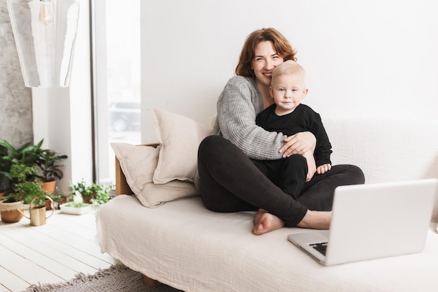 Jonge lachende vrouw zittend op de Bank met haar mooie zoontje gelukkig in de camera kijken Moeder en babyjongen vreugdevol tijd doorbrengen met laptop in gezellige woonkamer thuis