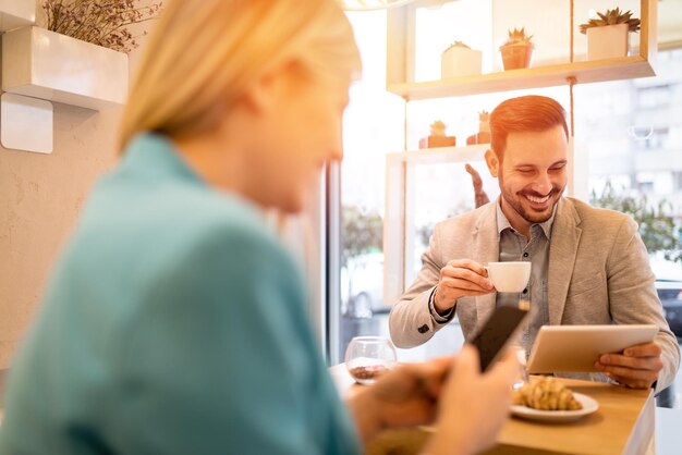 Jonge lachende zakenmensen op een pauze in een café. Man aan het werk op tablet en koffie drinken. Vrouw met behulp van slimme telefoon. Selectieve aandacht. Focus op de achtergrond, op zakenman.