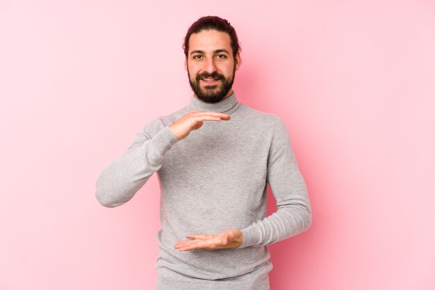 Jonge lange haarmens die op roze wordt geïsoleerd die iets met beide handen, productpresentatie houdt.