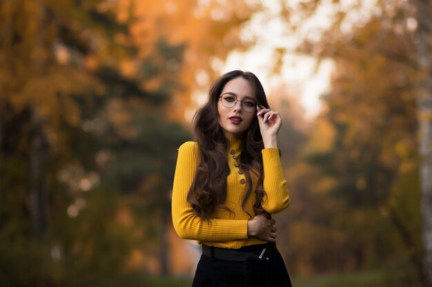 Jonge langharige brunette in gele gebreide trui en bril die naar de camera kijkt terwijl ze tegen wazig gebladerte van herfstbomen in het park staat