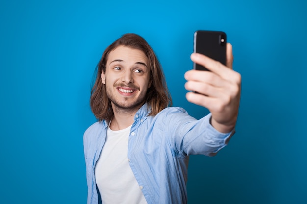 Jonge langharige man met baard lacht en maakt een selfie met behulp van een telefoon op een blauwe studiomuur in vrijetijdskleding