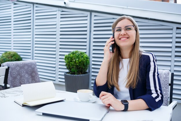 Jonge langharige vrouw praten op een mobiele telefoon, werken met een laptop, zittend aan een houten tafel op een terras in een café