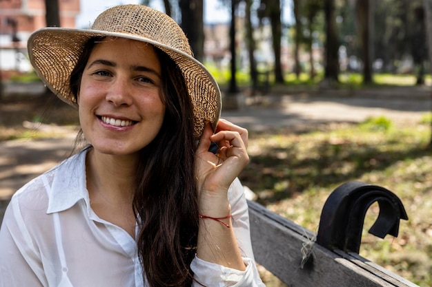 Jonge Latijns-Amerikaanse vrouw 33 zittend op een bankje in het park is blij en glimlachend bij de komst van de lente Ze draagt een hoed en een wit overhemd Concept van de lente