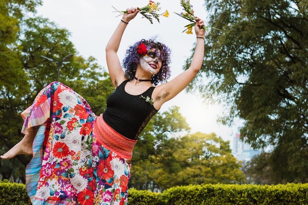 Jonge latijns-kaukasische vrouw die in het park danst met een kleurrijke jurk en catrina-make-up
