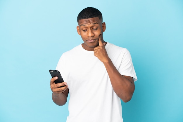 Jonge latijns-man geïsoleerd op blauwe muur met behulp van mobiele telefoon en denken