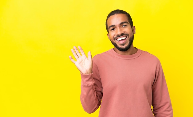 Jonge Latijns-man geïsoleerd op gele achtergrond saluerend met de hand met gelukkige uitdrukking