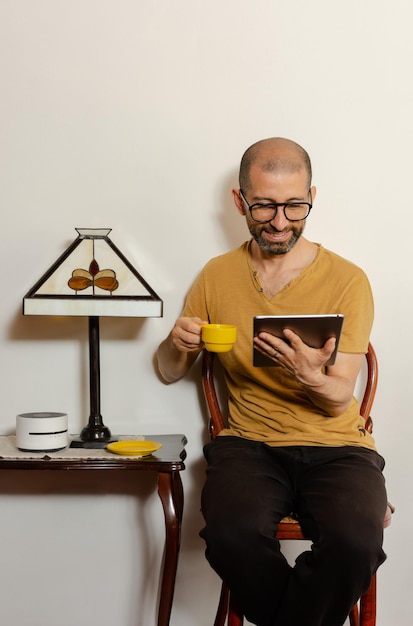 Jonge Latijns-man in vrijetijdskleding die op een tablet leest terwijl hij een kopje glimlachend thuis op een stoel vasthoudt Kopieer de ruimte