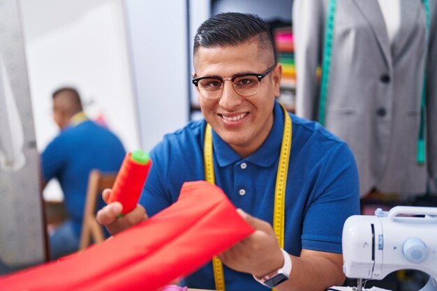 Jonge latijns-man kleermaker glimlachend zelfverzekerd met doek en draad in atelier