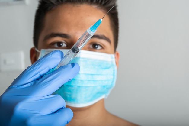Jonge Latijns-man met chirurgisch masker zonder shirt met spuit in de hand voorbereid op vaccinatie