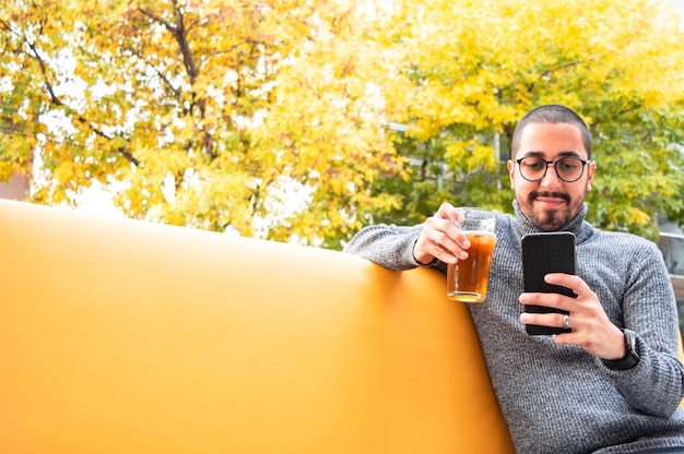 Jonge Latijns-man zit op een meubel buiten een bar bier te drinken en zijn telefoon