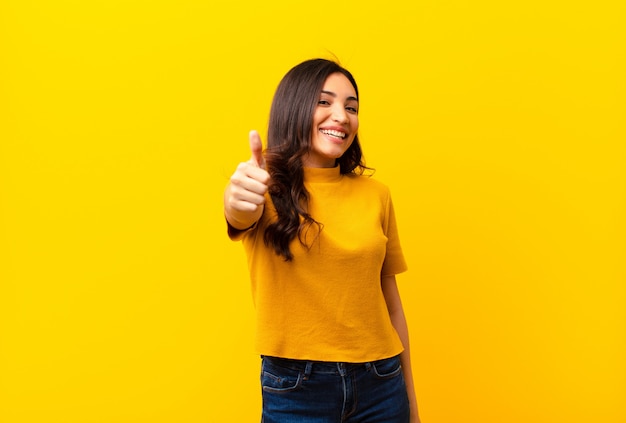 Jonge latijns-mooie vrouw voelt zich trots, zorgeloos, zelfverzekerd en gelukkig, glimlachend positief met duimen omhoog over muur