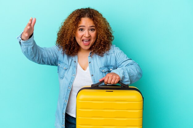 Jonge Latijns-reiziger bochtige vrouw met een koffer geïsoleerd op blauwe achtergrond die een aangename verrassing ontvangt, opgewonden en handen opsteekt.