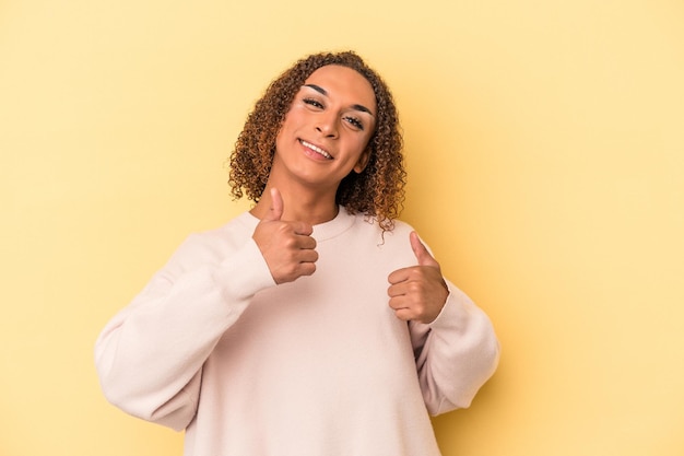 Jonge Latijns-transseksuele vrouw geïsoleerd op gele achtergrond die beide duimen opheft, glimlachend en zelfverzekerd.