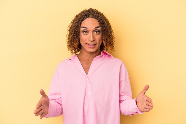 Jonge Latijns-transseksuele vrouw geïsoleerd op gele achtergrond met iets met beide handen, productpresentatie.