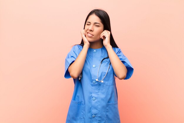 Jonge Latijns-verpleegster kijkt boos, gestrest en geïrriteerd en bedekt beide oren met een oorverdovend geluid, geluid of luide muziek tegen een roze muur
