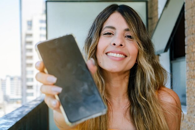 Jonge latijns-vrouw die lacht telefoon vasthouden met uitgestrekte hand voor haar