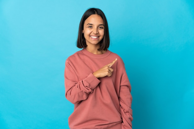 Jonge Latijns-vrouw die op blauwe achtergrond wordt geïsoleerd die naar de kant wijst om een product te presenteren