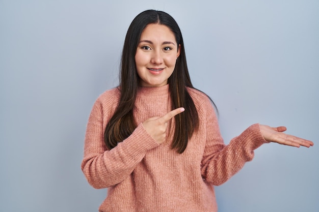 Jonge latijns-vrouw die over blauwe achtergrond staat verbaasd en glimlachend naar de camera terwijl ze met de hand presenteert en met de vinger wijst