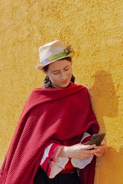 Jonge latijns-vrouw in boerenkleding leunend tegen een gele muur terwijl ze haar mobiele telefoon controleert