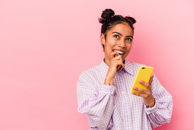 Jonge latijns-vrouw met een mobiele telefoon geïsoleerd op roze achtergrond ontspannen denken over iets kijken naar een kopie ruimte.