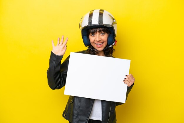 Foto jonge latijns-vrouw met een motorhelm geïsoleerd op gele achtergrond met een leeg bordje en saluting