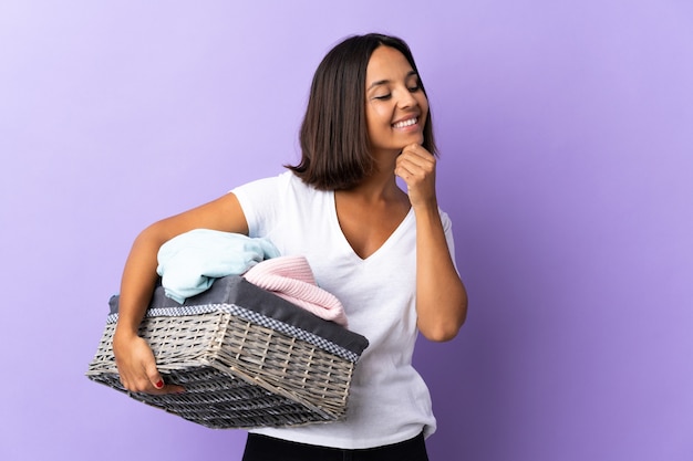 Jonge Latijns-vrouw met een wasmand geïsoleerd op paars op zoek naar de kant en glimlachen