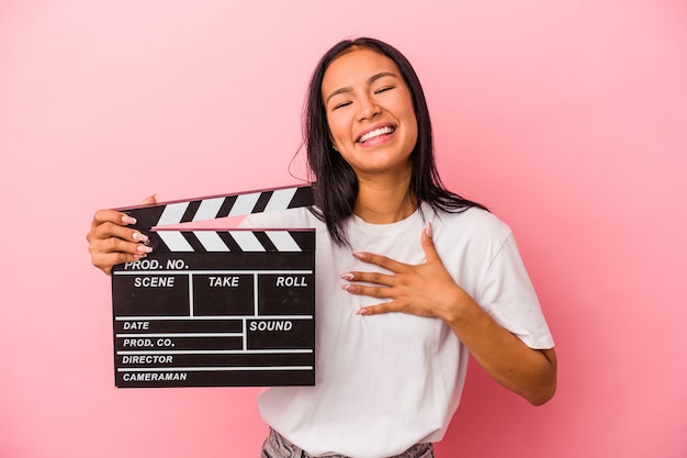 Jonge latijns-vrouw met filmklapper geïsoleerd op roze achtergrond lacht hardop terwijl ze de hand op de borst houdt.