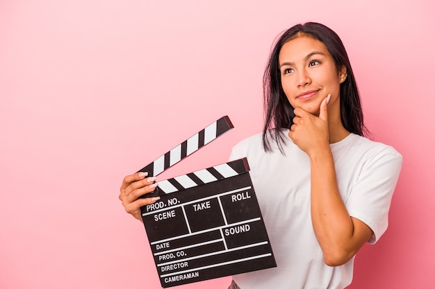 Jonge Latijns-vrouw met Filmklapper geïsoleerd op roze achtergrond zijwaarts kijkend met twijfelachtige en sceptische uitdrukking.