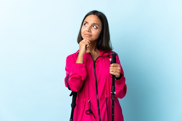 Jonge Latijns-vrouw met rugzak en wandelstokken geïsoleerd op blauw twijfels en denken