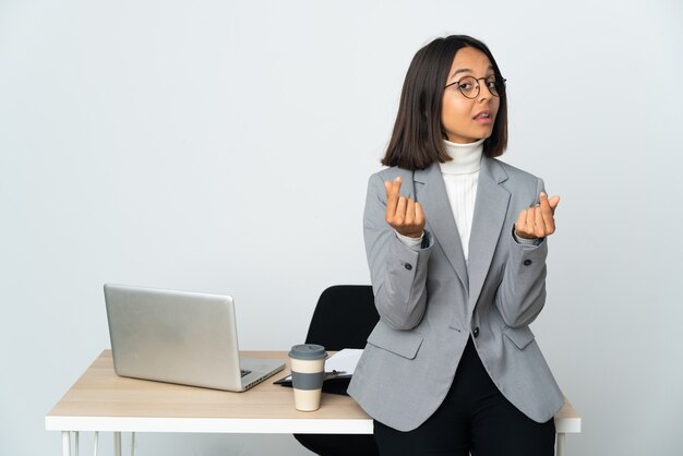 Jonge Latijnse bedrijfsvrouw die in een bureau werkt dat op wit wordt geïsoleerd dat geldgebaar maakt