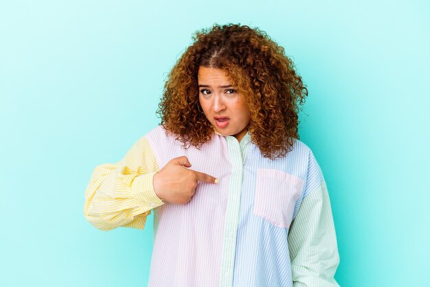 Jonge Latijnse bochtige vrouw die op blauwe muur wordt geïsoleerd verrast wijzend met vinger, breed glimlachend.