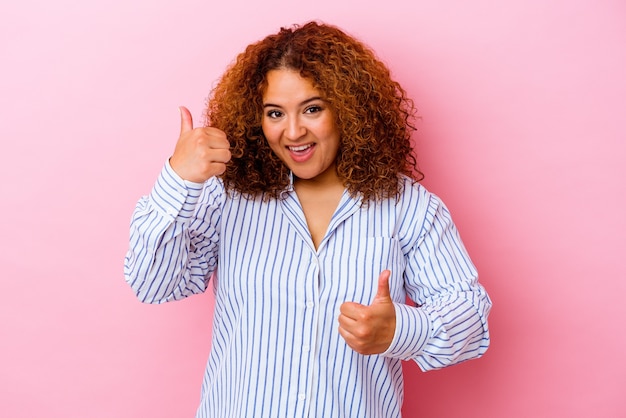 Jonge Latijnse bochtige vrouw die op roze achtergrond wordt geïsoleerd die beide duimen opheft, glimlachend en zelfverzekerd.