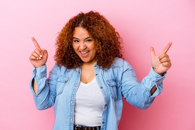 Jonge Latijnse bochtige vrouw die op roze muur wordt geïsoleerd die naar verschillende exemplaarruimten wijst, een van hen kiest, met vinger toont.