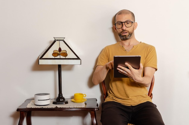 Jonge latijnse man in vrijetijdskleding met een tablet met een verbaasde uitdrukking die thuis op een stoel zit Kopieer de ruimte