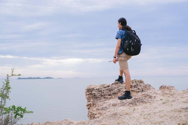 Jonge latijnse man met een rugzak die imposant op de rots van een klif voor het strand staat Kopieer de ruimte