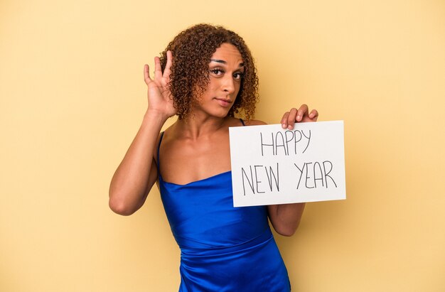 Jonge latijnse transseksuele vrouw die nieuwjaar viert geïsoleerd op gele achtergrond die een roddel probeert te luisteren.