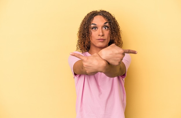 Jonge latijnse transseksuele vrouw geïsoleerd op gele achtergrond wijst zijwaarts, probeert te kiezen tussen twee opties.