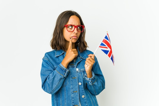 Jonge latijnse vrouw die een engelse vlag houdt die op witte muur wordt geïsoleerd die zijwaarts met twijfelachtige en sceptische uitdrukking kijkt.