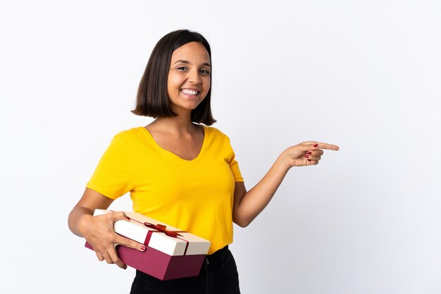 Jonge Latijnse vrouw die een gift houdt die op wit wordt geïsoleerd dat vinger aan de kant richt en een product voorstelt