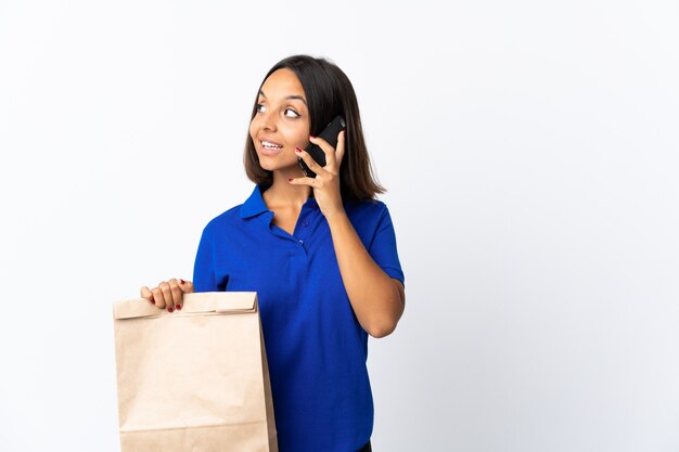 Jonge Latijnse vrouw die een kruidenierswinkel boodschappentas geïsoleerd op wit houden een gesprek met de mobiele telefoon met iemand