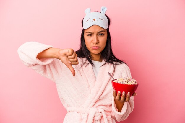 Jonge latijnse vrouw die een pyjama draagt met een kom granen geïsoleerd op een roze achtergrond met een afkeergebaar, duim omlaag. Onenigheid begrip.