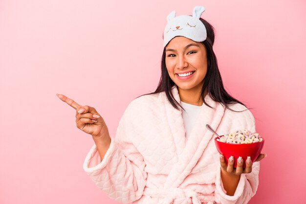 Jonge latijnse vrouw die een pyjama draagt met een kom ontbijtgranen geïsoleerd op een roze achtergrond glimlachend en opzij wijzend, iets tonend op de lege ruimte.