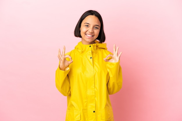 Jonge latijnse vrouw die een regenbestendige jas draagt over een geïsoleerde achtergrond met een ok teken met twee handen