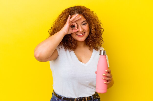 Jonge latijnse vrouw die een thermosfles houdt die op gele opgewekte achtergrond wordt geïsoleerd die ok gebaar op oog houdt.