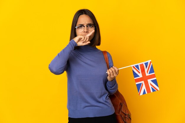 Jonge Latijnse vrouw die een vlag van het Verenigd Koninkrijk houdt die op gele achtergrond wordt geïsoleerd die stopgebaar met haar hand maakt om een handeling te stoppen