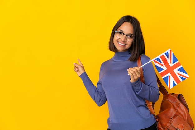 Jonge Latijnse vrouw die een vlag van het Verenigd Koninkrijk houdt die op gele achtergrond wordt geïsoleerd die terug richt